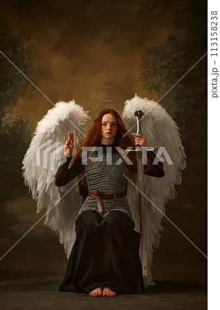 Redhead angel in chainmail, raising sword, with large white wings and golden halo against vintage studio background. Renaissance-style 113158238