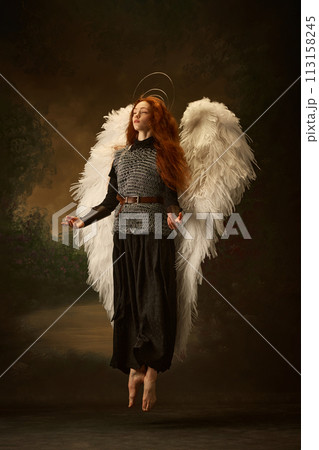 Serene redhead woman with white wings and halo, floating in old-fashioned chainmail top and black skirt against vintage studio background. Serene redhead woman with white wings and halo, floating in old-fashioned chainmail top and black skirt against vintage studio background. 113158245