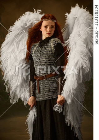Red-haired woman with angel wings and chainmail, wearing diadem, posing looking camera against vintage studio background. Red-haired woman with angel wings and chainmail, wearing diadem, posing looking camera against vintage studio background. 113158304