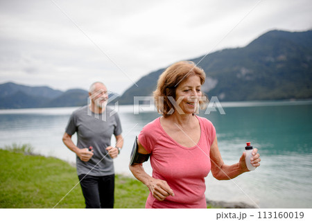 Senior couple running by lake in nature. Elderly husband and wife spending active vacation in the mountains, enjoying physical activity and relaxation outdoors. Senior couple running by lake in nature. Elderly husband and wife spending active vacation in the mountains, enjoying physical activity and relaxation outdoors. 113160019