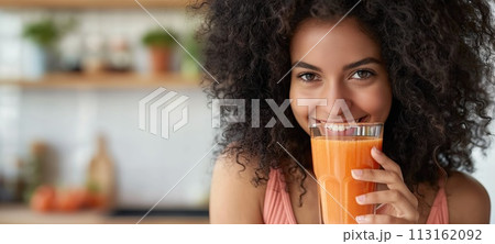 Portrait of a smiling young woman drinking carrot juice in the kitchen 113162092