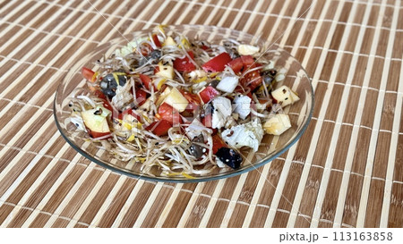Salad of fresh vegetables and microgreens. Tomatoes, onions, apples, a mound of sprouted mung beans, sesame seeds, flax and cheese cubes on on a transparent plate on the table. Close-up Salad of fresh vegetables and microgreens. Tomatoes, onions, apples, a mound of sprouted mung beans, sesame seeds, flax and cheese cubes on on a transparent plate on the table. Close-up 113163858