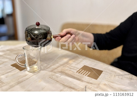 A woman's hand pours delicious dark tea from a glass teapot into a glass vessel standing on the table. Close-up A woman's hand pours delicious dark tea from a glass teapot into a glass vessel standing on the table. Close-up 113163912