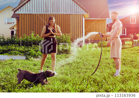 Playful dog grabs splashes of water from garden hose with its mouth during an evening walk in courtyard of country house with young European man and woman. Playful dog grabs splashes of water from garden hose with its mouth during an evening walk in courtyard of country house with young European man and woman. 113163936