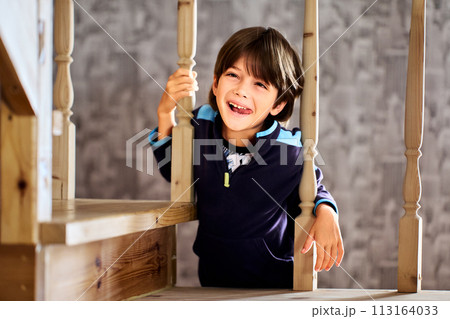 Seven year old boy stuck out his tongue while making faces on wooden stairs and holding onto balusters. 113164033
