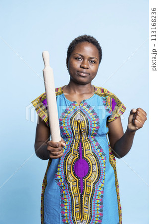 African american displeased person using rolling pin to threaten someone in front of camera, wearing traditional historic outfit. Young serious adult being aggressive, starting a fight. African american displeased person using rolling pin to threaten someone in front of camera, wearing traditional historic outfit. Young serious adult being aggressive, starting a fight. 113168236