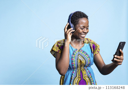 Charming happy woman dancing and listening to groovy music on headphones, using smartphone to change songs from a playlist. African american girl having fun with modern tunes in studio. Charming happy woman dancing and listening to groovy music on headphones, using smartphone to change songs from a playlist. African american girl having fun with modern tunes in studio. 113168248