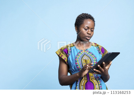 African american modern girl feels satisfied using a tablet over blue background, browsing online websites on mobile gadget. Young adult holds electronic device to navigate the internet. 113168253