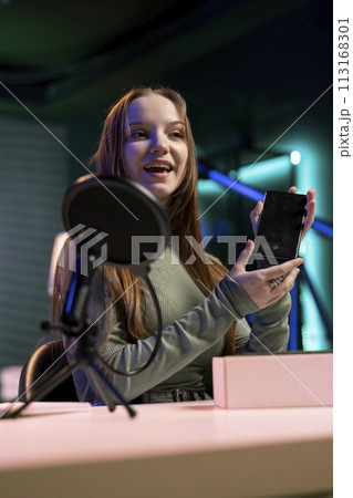 Teenager content creator filming technology review of newly released smartphone, unpacking it and presenting specifications to audience. Media star showing cellphone to fans from studio Teenager content creator filming technology review of newly released smartphone, unpacking it and presenting specifications to audience. Media star showing cellphone to fans from studio 113168301
