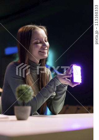 Happy young influencer in studio using camera to capture live broadcast, reviewing mini RGB lights. Joyful media star films electronics haul, presenting wireless LED lighting device Happy young influencer in studio using camera to capture live broadcast, reviewing mini RGB lights. Joyful media star films electronics haul, presenting wireless LED lighting device 113168333