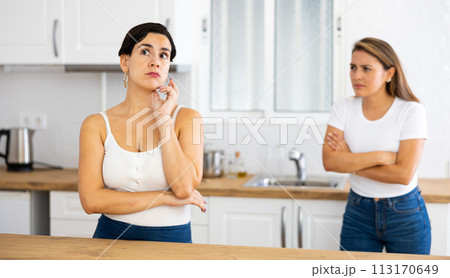 Offended Latina standing in kitchen, listening to disgruntled female friend Offended Latina standing in kitchen, listening to disgruntled female friend 113170649