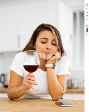 Frustrated young woman drinking wine and waiting for call in home kitchen Frustrated young woman drinking wine and waiting for call in home kitchen 113170673
