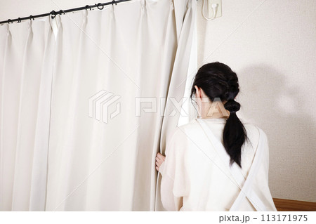 カーテンの隙間から外を見るエプロン姿の女性 113171975
