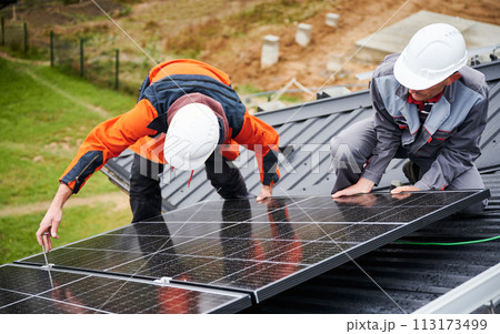 Mounters installing photovoltaic solar panels on roof of house. Men engineers in helmets building solar module system with help of hex key. Concept of alternative, renewable energy. 113173499