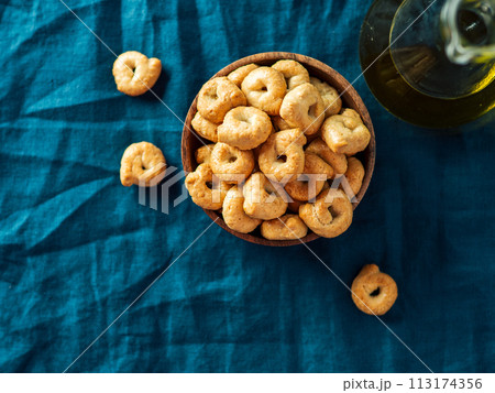 Traditional italian snack taralli or tarallini in wooden bowl over dark blue linen napkin background. Rustic shot of taralli appetizer with copy space. Top view or flat lay. 113174356