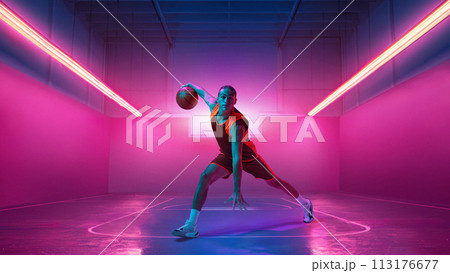 Dribbling ball. Young man in uniform basketball player in motion against gradient indoor stadium background in neon light. Dribbling ball. Young man in uniform basketball player in motion against gradient indoor stadium background in neon light. 113176677