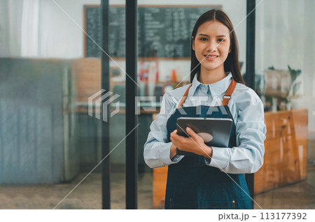Smiling Asian coffee shop owner standing in front of cashier counter with tablet ready to take menu orders.welcome customers who come in to place orders in the cafe. Smiling Asian coffee shop owner standing in front of cashier counter with tablet ready to take menu orders.welcome customers who come in to place orders in the cafe. 113177392