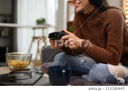 Happy smiling young woman wearing headset playing video game with joystick in living room Happy smiling young woman wearing headset playing video game with joystick in living room 113180445