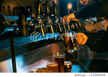 Hand drawing cold, refreshing, lager beer from tap with various other beers displayed at well-lit bar. Serving craft beer from tap. 113180594