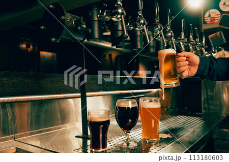 Photo of bartender pouring cold foamy beer from tap into glass at bar with multiple beer dispensers. Serving craft beer from tap. Photo of bartender pouring cold foamy beer from tap into glass at bar with multiple beer dispensers. Serving craft beer from tap. 113180603