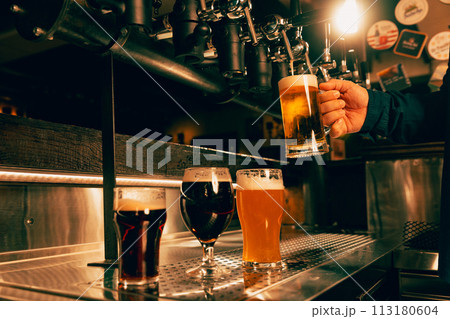 Photo of bartender's hand holds glass of light, cold, refreshing lager beer. Man Serving craft beer from tap at traditional pub. Photo of bartender's hand holds glass of light, cold, refreshing lager beer. Man Serving craft beer from tap at traditional pub. 113180604