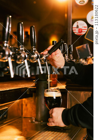 Bartender pouring dark, cold foamy beer from tap into glass at bar with multiple beer dispensers. Serving craft beer from tap. Oktoberfest concept. Bartender pouring dark, cold foamy beer from tap into glass at bar with multiple beer dispensers. Serving craft beer from tap. Oktoberfest concept. 113180622