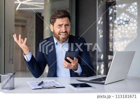 Portrait of an angry young businessman sitting at a desk in the office, holding a phone in his hand and looking disappointed at the camera. 113181671
