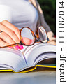 Woman's hand resting on an open book, holding reading glasses, close-up 113182034