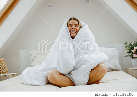 Happy woman is sitting on a bed wrapped in a white blanket Happy woman is sitting on a bed wrapped in a white blanket 113182100