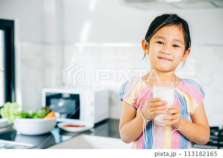 Cute Asian little girl in kitchen holds milk smiling joyfully. Portrait of daughter enjoying drink at home. Happy preschooler savoring calcium-rich liquid radiating happiness give me. Cute Asian little girl in kitchen holds milk smiling joyfully. Portrait of daughter enjoying drink at home. Happy preschooler savoring calcium-rich liquid radiating happiness give me. 113182165