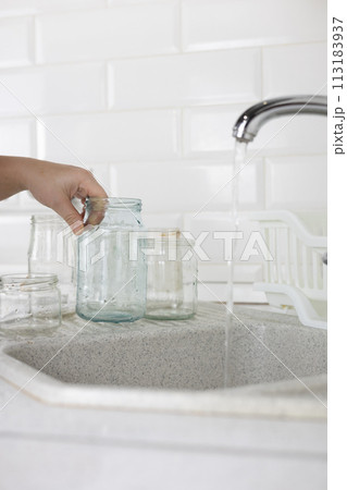 Vertical. Glass sorting. In the kitchen sink, a woman under the tap washes a glass jar to be sent for recycling. A woman sorts garbage to be sent for recycling and reuse. Eco activism concept. 113183937