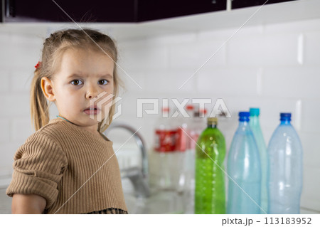 Pensive child sorts plastic in the kitchen and looks at the camera. The child actively helps the family sort plastic for recycling. The concept of caring for the planet. Pensive child sorts plastic in the kitchen and looks at the camera. The child actively helps the family sort plastic for recycling. The concept of caring for the planet. 113183952