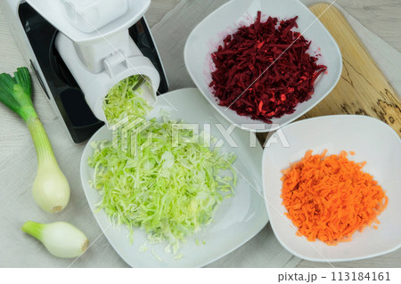 Cabbage, carrot, beets and onion in a vegetable cutter on kitchen table. Chopped carrot is falling into a bowl. Homemade healthy food. Health line. 113184161