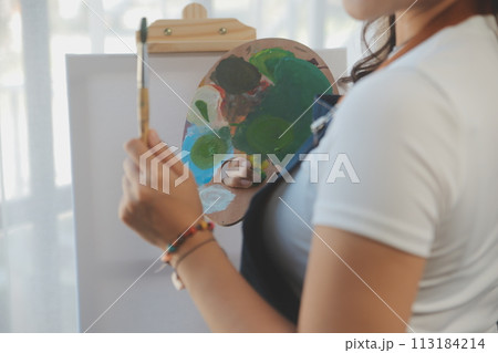 Cropped image of female artist standing in front of an easel and dipping brush into color palette 113184214