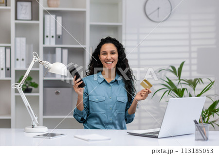 Happy Hispanic businesswoman with smartphone and credit card, working from a cozy modern home office, smiling confidently. 113185053