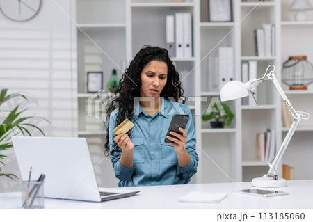 Concerned Hispanic woman reviewing credit card and smartphone, finding a mistake while working from her home office. 113185060