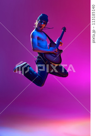 Energetic young shirtless man in sunglasses,rocket playing guitar against pink background in neon light 113185140
