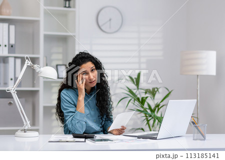 A Hispanic businesswoman appears confused while working in her home office, surrounded by her work essentials. 113185141