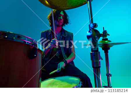 Artistic young boy in stylish clothes, musician playing drums against blue background in neon light. Rock and roll 113185214
