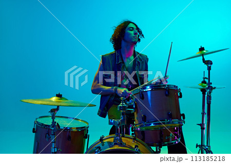Young boy, musician in casual clothes playing drums, training against blue background in neon light Young boy, musician in casual clothes playing drums, training against blue background in neon light 113185218