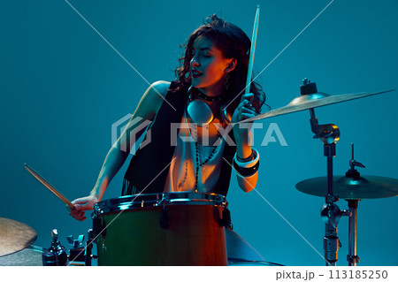 Beautiful young woman in stylish clothes, musician playing drums against cyan background in neon light. Rock and roll event 113185250