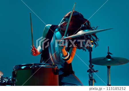 Beautiful, energetic young woman, artistic musician playing drums against cyan background in neon light 113185252