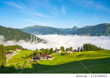 Morning clouds blanket the valley below a serene alpine landscape in Kitzbueheler Alps, with chalets and mountain peaks in view. Austria 113189401