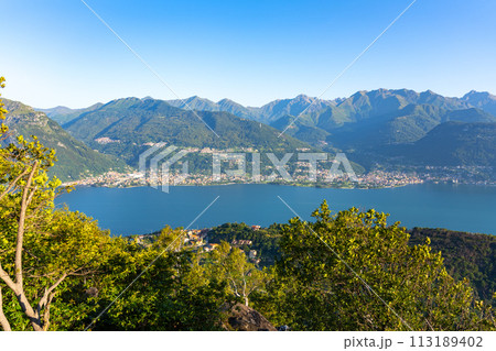 A beautiful mountain range with a lake in the foreground. The lake is calm and the mountains are covered in trees. The sky is clear and the sun is shining brightly. The scene is peaceful and serene 113189402