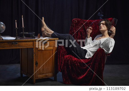 Young, thoughtful man in historical, old-fashion costume sitting in vintage chair drinking red wine and smoking cigarettes Young, thoughtful man in historical, old-fashion costume sitting in vintage chair drinking red wine and smoking cigarettes 113189702