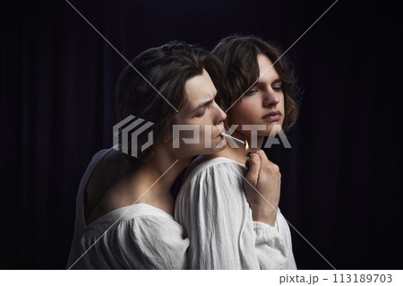 Two people, in white old-fashioned shirts, one lighting cigarette while hugs other against dark velvet background. Two people, in white old-fashioned shirts, one lighting cigarette while hugs other against dark velvet background. 113189703