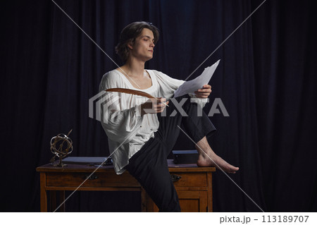 Young man, poet in historical costume sitting on vintage desk and writing poem with feather against dark background. 113189707