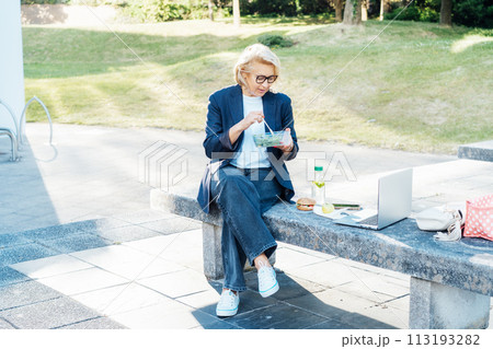 Business woman eating fast and healthy in front of laptop during job lunch break outdoors. Modern business lifestyle. Middle aged lady eats salad and use notebook pc on bench. Balanced diet lunch box. 113193282