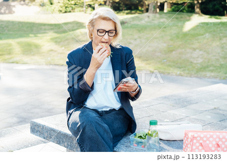 Middle-aged business woman eating sandwich burger with salad and using phone sitting on bench in the park while having lunch break. Remote work,business, freelance,blogging,social media concept 113193283
