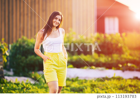 Yellow Bermuda shorts are worn by young European woman in countryside on summer evening, knee-length horizontal portrait. 113193783